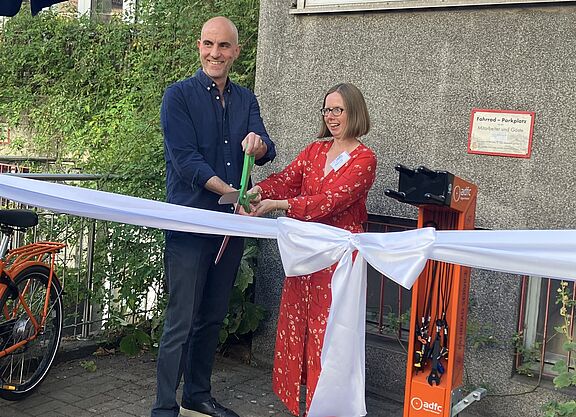 Ein Mann und eine Frau zershcneiden mit einer riesigen Schere ein weißes Band mit Schleife vor einer Fahrradreparatursäue